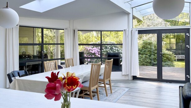 En matplats med ett bord, stolar, en vas med blommor och utsikt över trädgården genom stora fönster.