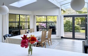 Een eethoek met een tafel, stoelen, een bloevas en uitzicht op de tuin door grote ramen.