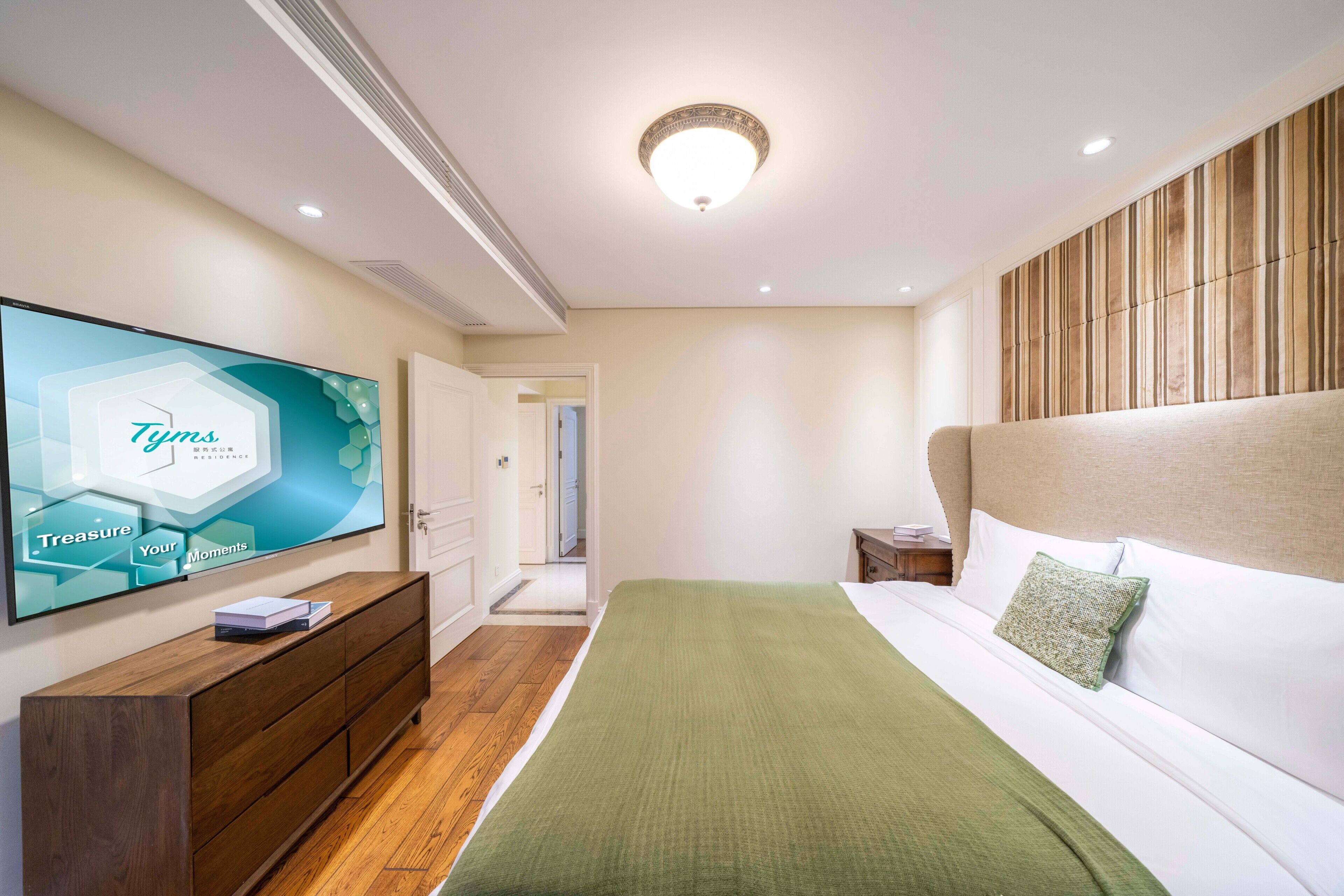 A modern hotel room with a large flat-screen TV, a wooden dresser, a bed with a green blanket, and a wooden headboard.
