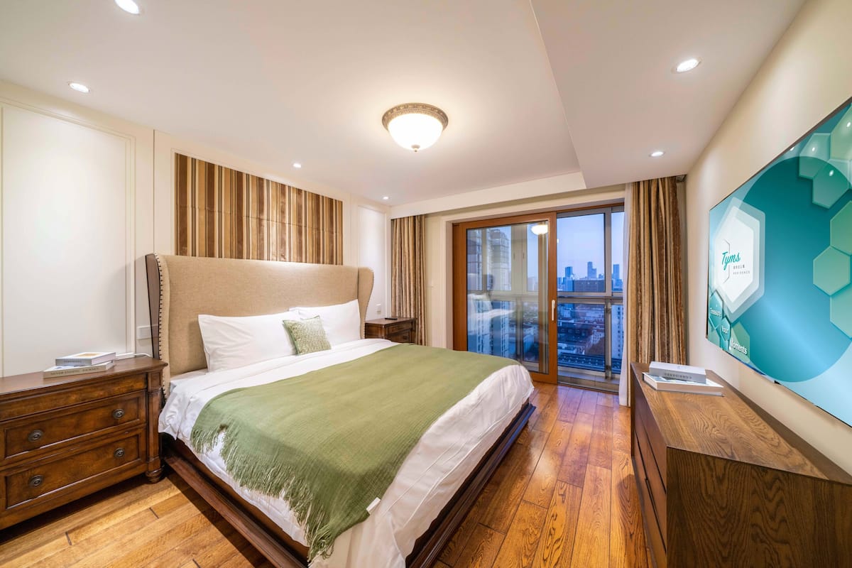 Quarto de hotel com uma cama grande, uma cômoda de madeira, uma televisão e uma varanda com vista para a cidade.
