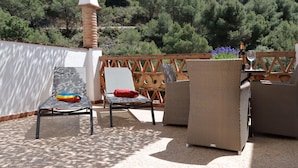 Um pátio externo com mobiliário de vime, uma mesa com garrafa e copo, e vista para uma encosta arborizada.