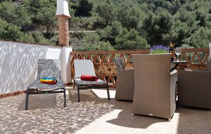 Eine Terrasse im Freien mit Korbmöbeln, einem Tisch mit Flasche und Glas sowie Blick auf einen bewaldeten Hang.