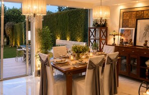 A dining area with a table set for four, surrounded by chairs, with a view of a garden and a hedge. - 3 bedroom stunning home in Benalmádena (Benalmádena)