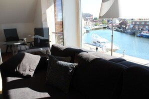 Ein modernes Wohnzimmer mit einer Couch, einem kleinen Tisch und einem Laptop. Ein großes Fenster bietet Blick auf einen Kanal mit Booten und Gebäuden.