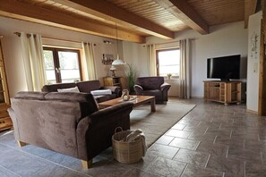 Ein Wohnzimmer mit Sofa, Sesseln, Couchtisch, TV-Schrank und einem großen Fenster.