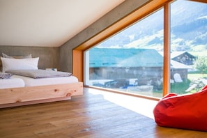 Ein Schlafzimmer mit Holzboden, einem Bett mit weißer Bettwäsche, einem großen Fenster mit Blick auf Berge und Gebäude sowie einem roten Sitzsack.