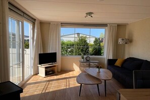 A living room with a TV, a sofa, a table, and a window with a view of greenery and buildings. - Cabin in Renesse with swimming pool (Renesse)