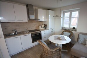 A modern kitchen with white cabinets, a stainless steel sink, and a built-in oven. There is a round dining table with wicker chairs. - Holiday apartment with 2 bedrooms (Sankt Peter-Ording)