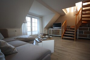 A loft-style room with a sofa, a television on a stand, a wooden staircase, and a window with curtains. - Holiday apartment with 2 bedrooms (Sankt Peter-Ording)