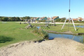 Ein Spielplatz mit Schaukeln und Rutschen, ein kleiner Teich und Häuser im Hintergrund.
