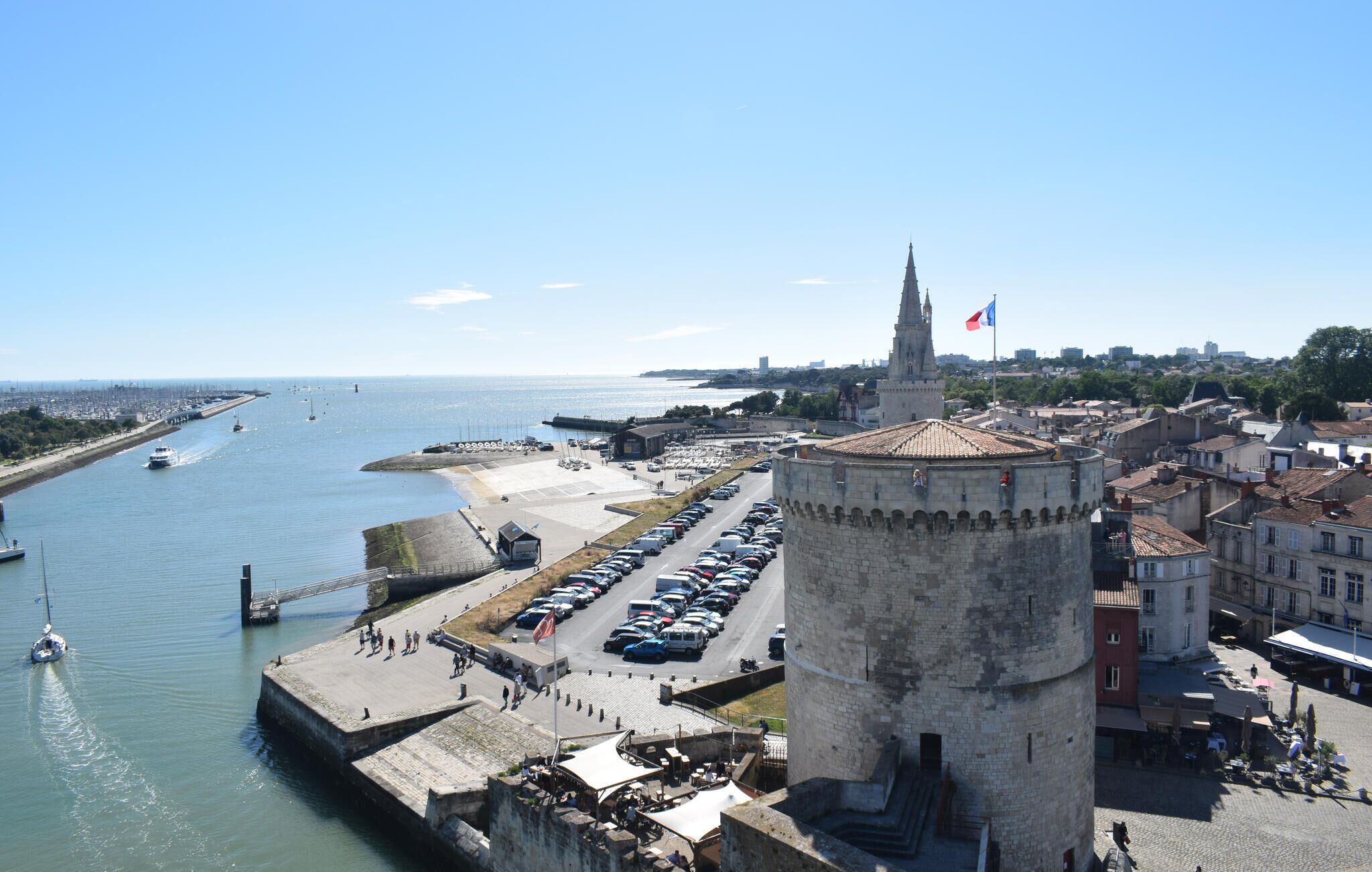 A coastal cityscape with a prominent tower, a marina with boats, and a clear view of the sea.