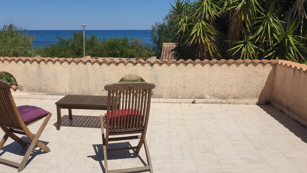 Un patio al aire libre con dos sillas de madera y una mesa, piso de baldosas y vista al mar.