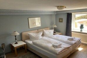 Ein Doppelbett mit weißen Leinentüchern, zwei Nachttische mit Lampen, ein gerahmtes Bild an der Wand und ein Fenster mit Vorhängen.