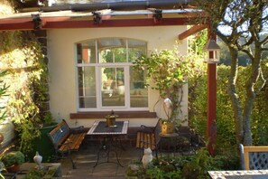 Eine überdachte Terrasse mit Tisch und Bänken, umgeben von Pflanzen und einem Fenster.