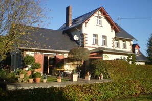 Ein zweistöckiges Haus mit geneigtem Dach, Schornstein und einer Veranda mit Blumentöpfen.