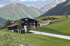 Eine Berghütte mit einer daranführenden Straße, umgeben von grünen Hügeln und schneebedeckten Bergen.