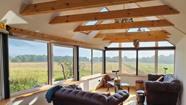 A spacious room with large windows, a brown leather sofa, a small table, and a chair. - Cosy Cottage close to Munros and cycle route (Lochearnhead)