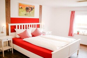 A bed with a red and white bedspread, a white nightstand, a red and white checkered curtain, and a painting of a cow on the wall. - Holiday home in Hayingen with barbecue (Hayingen)