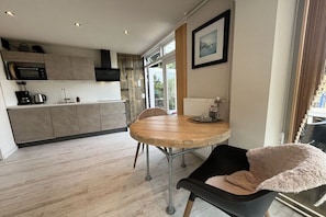A modern kitchen with a round wooden table, a black chair with a fluffy cushion, and a view of the outdoors through a large window. - Holiday home with 1 bedroom (Callantsoog)
