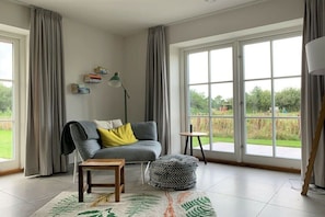 Ein geräumiges Zimmer mit grauem Sofa, einem kleinen Holztisch, einem Sitzsack und einem großen Fenster mit Blick auf Grünflächen.
