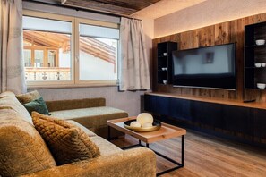 Ein Wohnzimmer mit einer Couch, einem Couchtisch mit einer Dekoration, einem an einer Holzvertäfelung montierten Fernseher und einem Fenster mit Vorhängen.