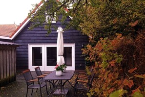 Eine Terrasse mit Tisch und Stühlen, einem weißen Sonnenschirm und einem Haus mit dunkler Fassade.