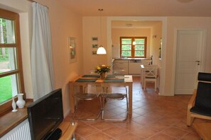 Ein Wohnzimmer mit Fernseher, zwei Sesseln, einem Esstisch mit Blumenvase und einer Küche im Hintergrund.