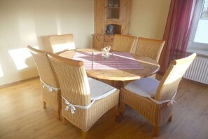 Ein runder Holztisch mit sechs Korbstühlen, einer gestreiften Tischdecke und einem Schrank mit Glastüren im Hintergrund.