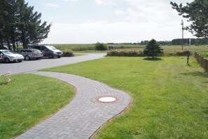 A paved pathway leading to a parking area with cars, surrounded by grass and trees. - Hüs Lennies DG South (Nebel \/ Amrum)