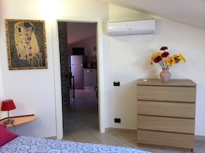 A bedroom with a bed, a dresser, a painting, a vase with flowers, and a kitchen in the background. - Apartment with terrace within walking distance of the historic center (Oristano)