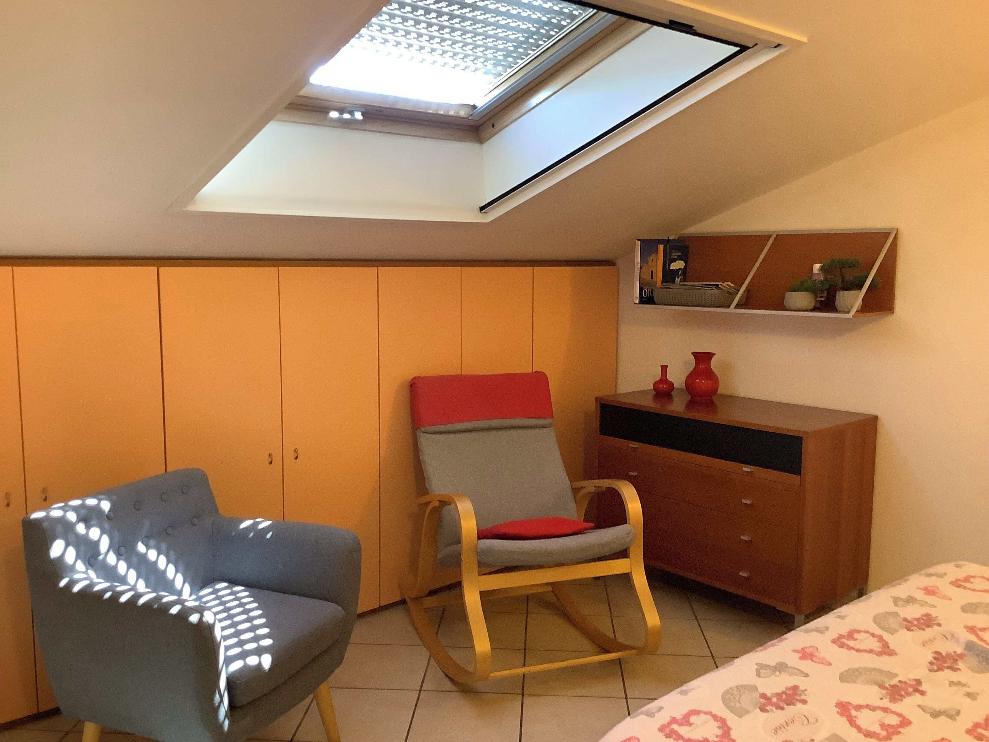 Una camera da letto con un lucernario, un letto con biancheria a motivi, una poltrona grigia, una sedia a dondolo, un comò in legno e uno scaffale con oggetti decorativi.