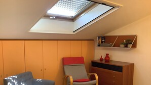 Ein Schlafzimmer mit Dachfenster, einem Bett mit gemusterten Betttextilien, einem grauen Sessel, einem Schaukelstuhl, einer Holzkommode und einem Regal mit Dekoration.