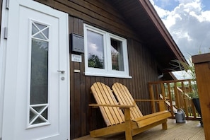 Ein Holzhaus mit einer weißen Tür und Fenstern, eine Holzterrasse mit zwei Adirondack-Stühlen und einem Schild mit der Aufschrift 'Casa 38b'.