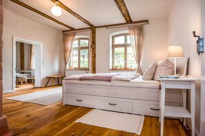 Ein Schlafzimmer mit Holzboden, einem Bett mit weißem Kopfteil, zwei Vorhängen an den Fenstern, einem Nachttisch mit Lampe sowie einem kleinen Tisch und einem Stuhl in der Ecke.