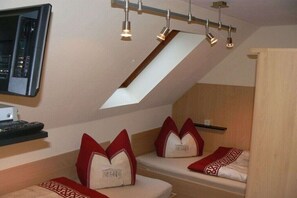 Ein Zimmer mit schrägem Dach, einem Bett mit roten und weißen Kissen, einem kleinen Regal mit einer Tasse und einem angebrachten Fernseher.