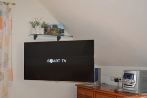 Ein Zimmer mit einem an der Wand befestigten Flachbildfernseher, einer kleinen Ablage mit Dekoration, einem Holzschrank und einem Drucker auf einem Schreibtisch.