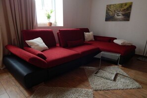 Un canapé d’angle rouge, une petite table d’appoint, une plante en pot près de la fenêtre et un tableau accroché au mur.