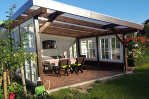 Une terrasse couverte avec une table et des chaises, un sol en briques et une vue sur une maison dotée de grandes fenêtres.