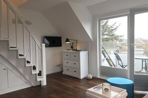 A modern room with a staircase, a flat-screen TV, a desk with drawers, and a view through a large window. - Holiday apartment with 2 bedrooms (Westerland)