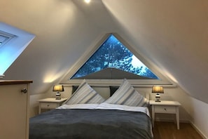 A bedroom with a sloped ceiling, a skylight, a bed with striped bedding, two bedside tables with lamps, and a view of trees through the skylight. - Holiday apartment with 2 bedrooms (Westerland)