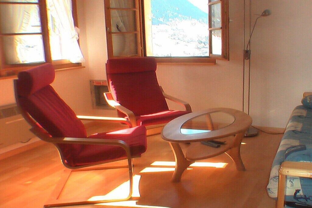 Ein Zimmer mit zwei roten Stühlen, einem runden Couchtisch, einer Couch und einem Fenster mit Blick auf eine verschneite Landschaft.