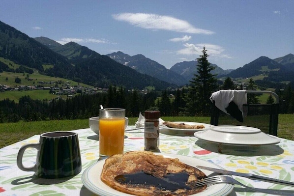 A breakfast table with a plate of pancakes, a mug, a glass of orange juice, and a bottle of syrup, set against a mountain backdrop. - Holiday apartment with 2 bedrooms (Riezlern)
