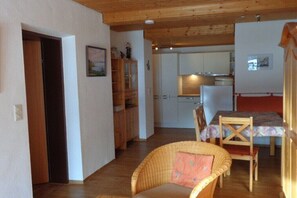 A room with a dining table, chairs, a wicker chair, a kitchen area with cabinets, and a refrigerator. - Holiday apartment with 2 bedrooms (Riezlern)