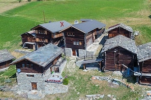 Eine Gruppe Holzhütten mit Stein Fundamenten, ein auffälliges rotes 'X' auf einem Dach und eine Satellitenschüssel an einem Gebäude.