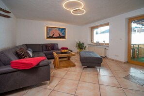 Ein modernes Wohnzimmer mit einem grauen Sofa, einem Holztisch und einem grauen Hocker. An der Wand hängt ein Bild, ein Fenster bietet einen Ausblick und eine Topfpflanze steht dort.