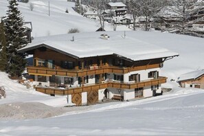 Ein mehrgeschossiges Chalet mit verschneitem Dach, Holzbalkonen und einer Skipiste im Hintergrund.
