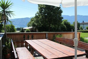 Ein Holztisch und eine Bank unter einem weißen Sonnenschirm, mit Blick auf Berge und ein Haus im Hintergrund.