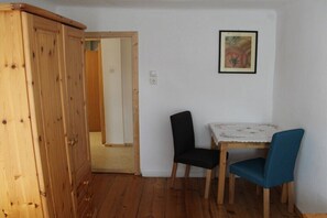Ein Zimmer mit einem Holzschrank, einem Tisch mit zwei Stühlen, einem gerahmten Bild an der Wand und einer Tür zu einem weiteren Raum.