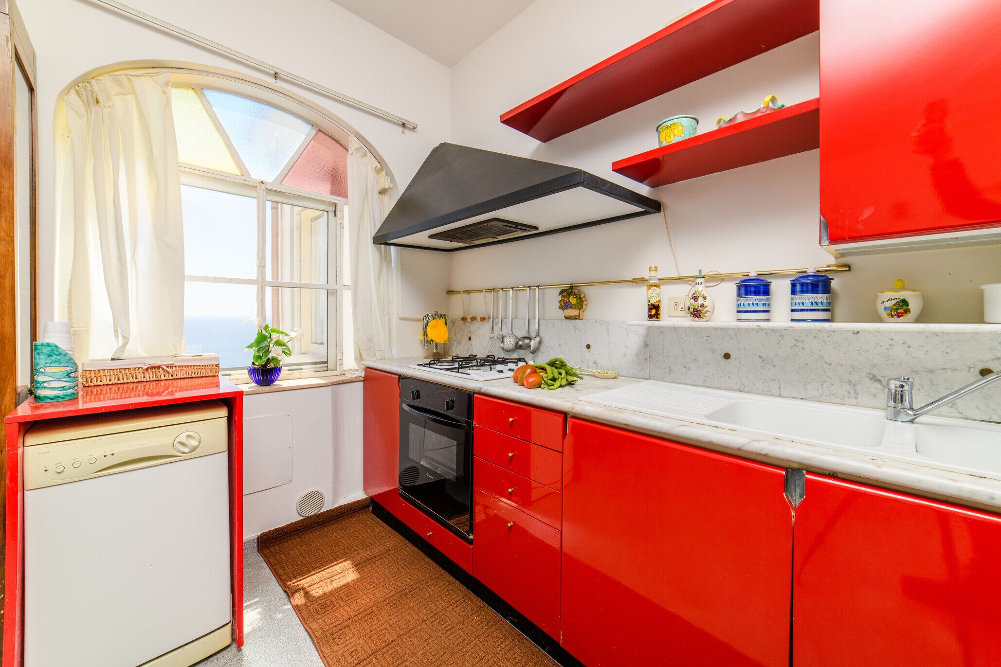 A kitchen with red cabinets, a white countertop, a stove, and a window with curtains.