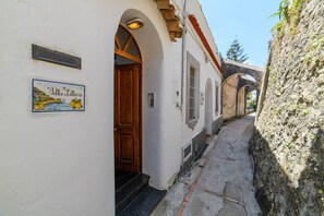 A narrow alleyway with a white building, a wooden door, and a sign with a coastal scene. - Estate4home - Villa Lettieri (Praiano)